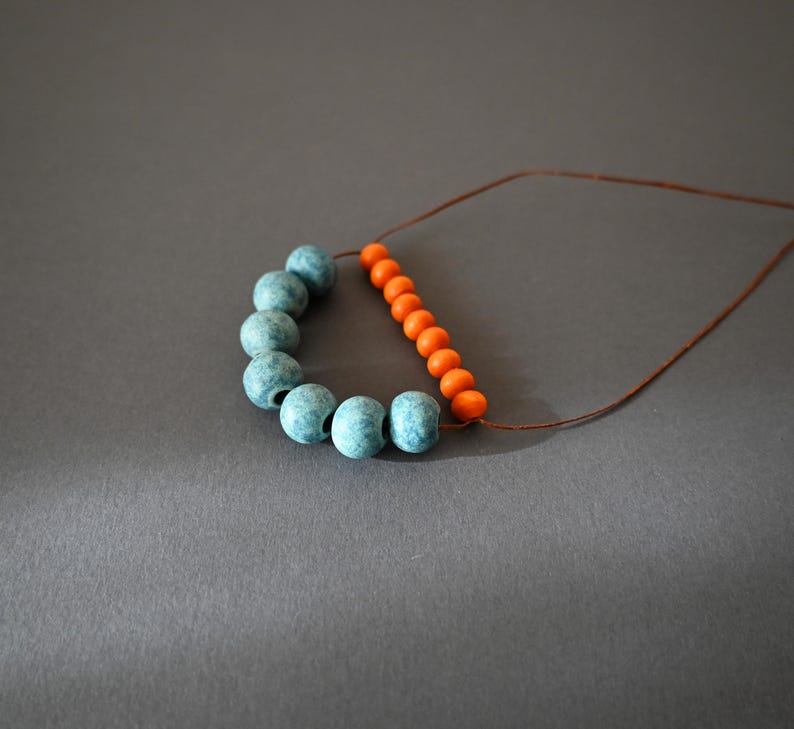 May include: A necklace featuring a combination of blue and orange beads. The blue beads are clustered in a curved line, while the orange beads form a straight line. The beads are strung on a thin, brown cord, creating a simple yet striking design.