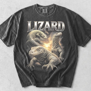 Può includere: Una t-shirt grigio antracite a maniche corte con una stampa grafica di tre lucertole. La parola LIZARD è stampata in grandi lettere dai toni metallici sopra i rettili, che si stagliano su uno sfondo di cielo nuvoloso al tramonto.