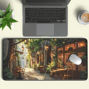 Puede incluir: Un ratón de ordenador y un portátil descansan sobre una alfombrilla de ratón rectangular grande. La alfombrilla muestra una ilustración detallada de una escena de calle europea con cafés, mesas y faroles colgantes. También se ven una pequeña planta en maceta y una taza de café.