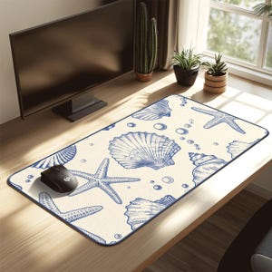 Può includere: Un tappetino per mouse rettangolare con sfondo color crema e illustrazioni blu di conchiglie e stelle marine. Un mouse per computer nero è appoggiato sopra. Il tappetino ha un bordo blu scuro.
