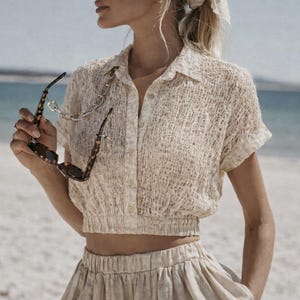 May include: A woman wearing a beige, textured, short-sleeved shirt with a collared neckline and matching skirt. She is holding tortoiseshell sunglasses with a gold chain. The outfit is light-colored and suitable for warm weather.
