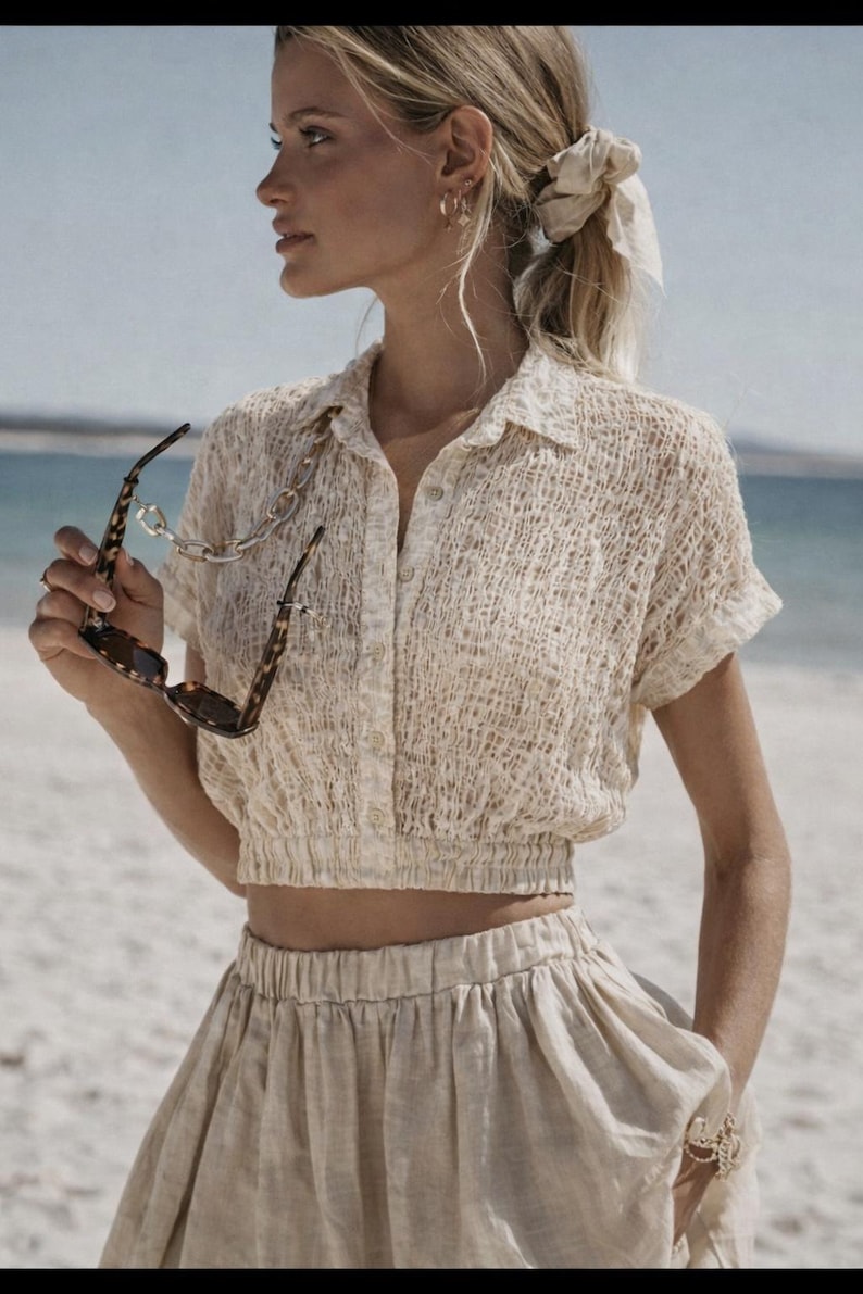 May include: A woman wearing a beige, textured, short-sleeved shirt with a collared neckline and matching skirt. She is holding tortoiseshell sunglasses with a gold chain. The outfit is light-colored and suitable for warm weather.