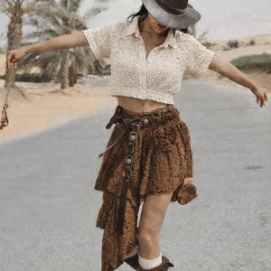 May include: A cream-colored crochet top and a brown skirt with a layered, asymmetrical design. The outfit is accessorized with a brown hat, belt, and boots. The background features palm trees and a road.
