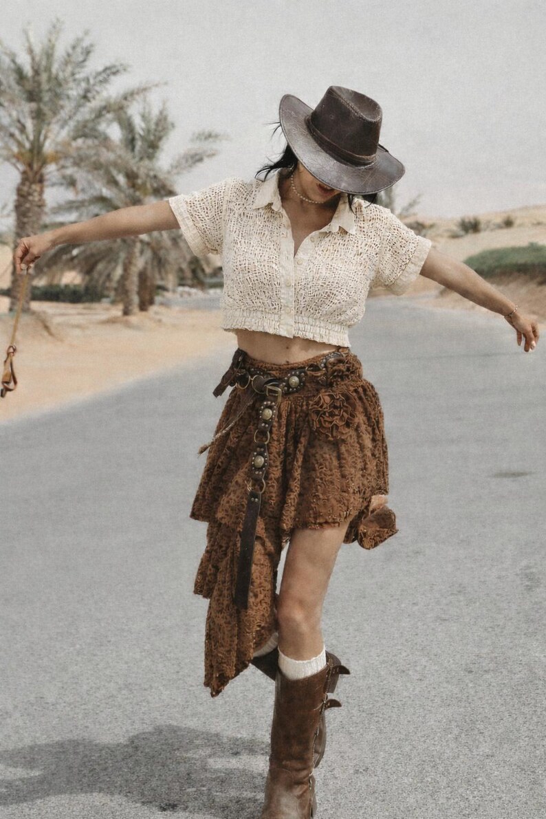 May include: A cream-colored crochet top and a brown skirt with a layered, asymmetrical design. The outfit is accessorized with a brown hat, belt, and boots. The background features palm trees and a road.