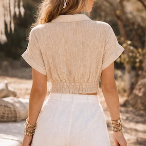 May include: A beige collared crop top with short sleeves and a cinched waist paired with white shorts. The outfit is accessorized with gold bracelets and a decorative headband. The style is bohemian.