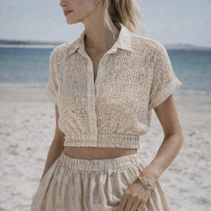 May include: A beige, short-sleeved crop top and matching skirt. The top has a collared neckline and button-down front. The skirt is a flowing, midi-length design. The outfit is made of a textured fabric, perfect for a beach day.