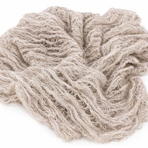 May include: A beige, loosely woven fabric, possibly linen or a similar material, is arranged in a soft, flowing pattern. The fabric has a textured appearance with visible gaps and a slightly distressed look, suitable for photography backdrops or craft projects.