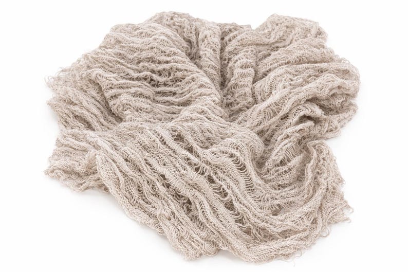 May include: A beige, loosely woven fabric, possibly linen or a similar material, is arranged in a soft, flowing pattern. The fabric has a textured appearance with visible gaps and a slightly distressed look, suitable for photography backdrops or craft projects.