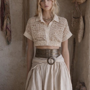 May include: A cream-colored outfit featuring a short-sleeved, open-weave top and a flowing skirt. A wide, ornate belt with a large circular buckle cinches the waist. The ensemble is accessorized with layered gold necklaces.