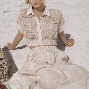 May include: A woman wearing a beige, open-weave short-sleeved shirt with a collared neckline and a matching skirt. The outfit is light-colored and appears to be made of a textured fabric. A picnic basket and food are visible in the background.