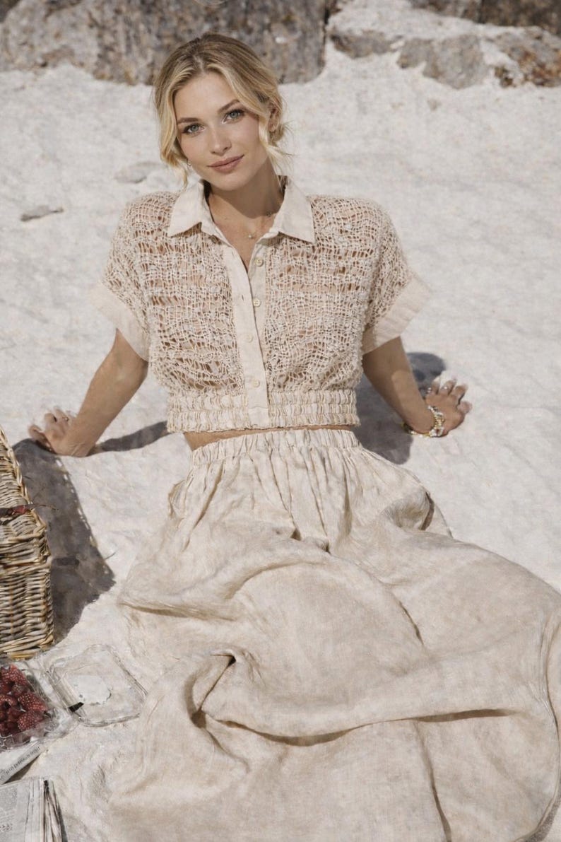 May include: A woman wearing a beige, open-weave short-sleeved shirt with a collared neckline and a matching skirt. The outfit is light-colored and appears to be made of a textured fabric. A picnic basket and food are visible in the background.