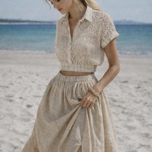 May include: A beige linen two-piece outfit, consisting of a short-sleeved, button-down crop top and a high-waisted maxi skirt with a side slit. The outfit is worn on a sandy beach with the ocean in the background.