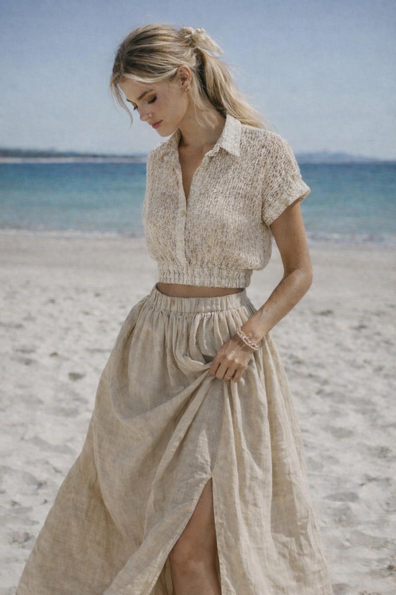 May include: A beige linen two-piece outfit, consisting of a short-sleeved, button-down crop top and a high-waisted maxi skirt with a side slit. The outfit is worn on a sandy beach with the ocean in the background.