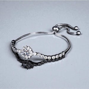 Puede incluir: Una pulsera plateada con un diseño floral central, incrustada con piedras preciosas transparentes. La pulsera presenta una delicada cadena y un cierre deslizante para un ajuste regulable. La banda es negra con piedras blancas.