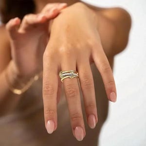 Può includere: Un anello dorato con un design intrecciato unico, caratterizzato da una fila di piccole pietre scintillanti. L'anello è mostrato su una mano con unghie curate, mettendo in risalto i suoi dettagli eleganti e intricati. Il design dell'anello è accattivante e adatto per l'uso quotidiano o occasioni speciali.