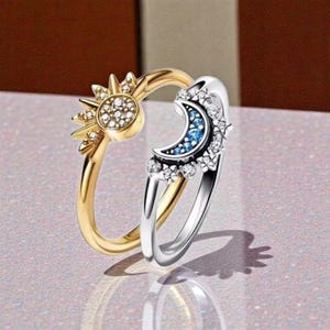 Può includere: Un anello a tema celeste con un design a sole dorato e luna argentata. Il sole ha una superficie strutturata con pietre trasparenti, mentre la luna ha pietre blu e pietre trasparenti circostanti. Un gioiello unico.