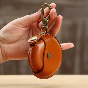 Könnte beinhalten: Ein runder, brauner Leder-Schlüsseletui mit einem Messingkarabiner und einem Knopfverschluss. Das Etui hat eine warme, mittelbraune Farbe und scheint aus hochwertigem Leder gefertigt zu sein. Der Verschluss ist bronzefarben.