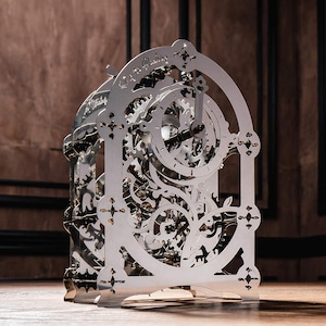 Op de afbeelding: Een zilverkleurige, ingewikkelde klok met zichtbare tandwielen en een decoratief frame. De klok heeft een ronde wijzerplaat met wijzers die de tijd aangeven. Het ontwerp omvat sierlijke uitsparingen en een gedetailleerd mechanisch interieur.