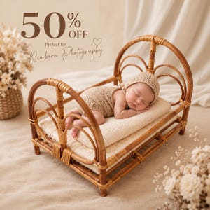 May include: A small, arched rattan bed frame with a light brown finish, featuring a woven base and curved side supports. A baby wearing a beige knit romper and matching bonnet is sleeping on a cream-coloured rectangular cushion inside the bed.
