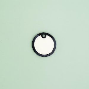 Könnte beinhalten: Ein runder, leerer weißer Anhänger mit schwarzem Rand und einem kleinen Loch oben zur Befestigung. Der Anhänger ist vor einem hellgrünen Hintergrund platziert.