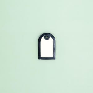 Puede incluir: Una etiqueta negra con forma de arco, interior blanco y un pequeño agujero en la parte superior. La etiqueta tiene un borde texturizado y está sobre un fondo verde claro. El diseño de la etiqueta es simple y elegante.