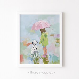 May include: A framed art print depicts a girl in a green raincoat and a pink umbrella, standing with a Dalmatian dog in the rain. The artwork has a soft, painterly style with pastel colors, creating a whimsical and heartwarming scene. The print is signed with the text "+ Hues & Hearth +" at the bottom.
