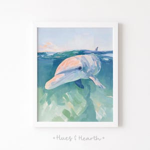 May include: A framed art print featuring a dolphin swimming underwater. The dolphin has a light pink and gray body with blue fins. The water is a mix of blue and green, with a hint of the sky above. The text "+ Hues & Hearth +" is at the bottom.