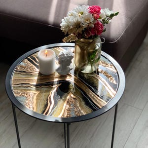 Op de afbeelding: Een ronde bijzettafel met een zwart metalen frame en een decoratief blad met gouden, zwarte en witte patronen. Een brandende witte kaars, een kleine buste en een glazen pot met bloemen staan op de tafel.