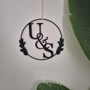 Könnte beinhalten: Schwarze Metall-Wanddekoration mit den Buchstaben "U & S" in einem Kreis, verziert mit Blättermotiven. Sie hängt an einer dünnen, naturfarbenen Schnur vor einer weißen Wand. Grüne Blätter sind im Hintergrund zu sehen.