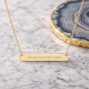 May include: Gold-colored bar necklace with the engraved text: "YEAH, WE CALL THIS SHIT NORMAL." The necklace hangs on a delicate chain. The background includes a marble surface and a decorative agate slice.