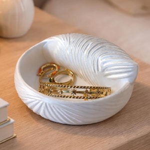 Puede incluir: Un joyero de cerámica blanca con diseño de pluma contiene anillos dorados y una pulsera. El plato está sobre una mesita de noche de madera junto a una lámpara y un pequeño jarrón con flores secas. El tema es la decoración del hogar.