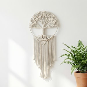 Puede incluir: Un colgante de pared de macramé beige con un diseño de árbol dentro de un marco circular. El árbol tiene ramas detalladas y un tronco texturizado. Debajo del árbol hay una serie de cuerdas colgantes, creando un efecto en capas. El colgante se muestra contra una pared blanca.