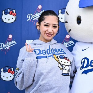 Op de afbeelding: Een grijze hoodie met het woord "Dodgers" in blauw over de borst. De hoodie heeft een Hello Kitty-afbeelding met een honkbalknuppel. De achtergrond is een blauwe muur met Hello Kitty- en Dodgers-logo's.
