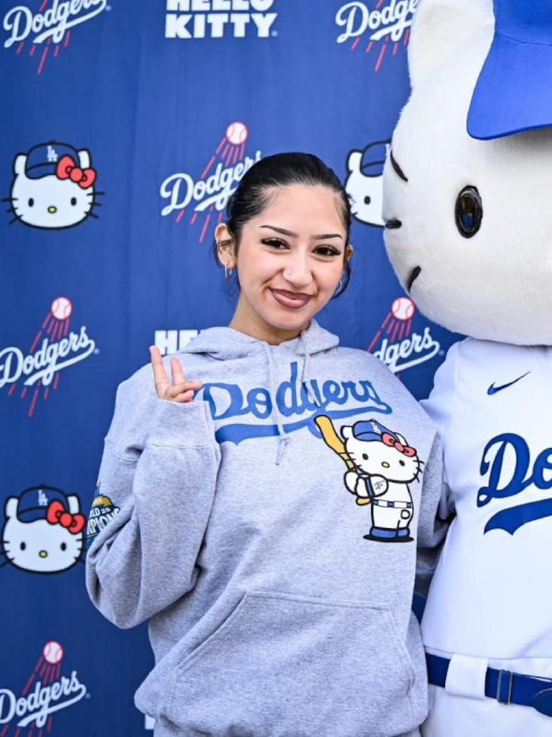 Op de afbeelding: Een grijze hoodie met het woord "Dodgers" in blauw over de borst. De hoodie heeft een Hello Kitty-afbeelding met een honkbalknuppel. De achtergrond is een blauwe muur met Hello Kitty- en Dodgers-logo's.