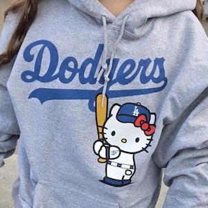 Op de afbeelding: Een grijze hoodie met het woord "Dodgers" in blauwe letters. De hoodie heeft een voorzak en een afbeelding van Hello Kitty met een Dodgers honkbalpet en een knuppel. De mouw heeft de tekst "Hello Kitty".