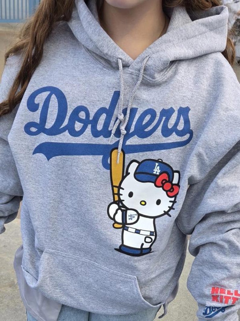 Op de afbeelding: Een grijze hoodie met het woord "Dodgers" in blauwe letters. De hoodie heeft een voorzak en een afbeelding van Hello Kitty met een Dodgers honkbalpet en een knuppel. De mouw heeft de tekst "Hello Kitty".