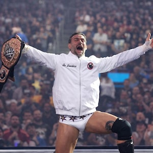 May include: A wrestler in a white jacket with "Bo Lueders" printed on the chest, and white trunks with star designs. He is holding a championship belt with both arms raised in celebration. The background is a blurred crowd.