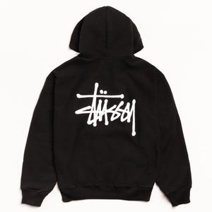 Op de afbeelding: Zwarte hoodie met een wit logo op de rug. Het logo is een gestileerd schrift met een kruisachtig symbool erboven. De hoodie heeft een capuchon met trekkoord en lange mouwen. Het kledingstuk is gemaakt van een zacht materiaal.