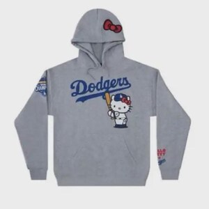 Op de afbeelding: Grijze hoodie met het woord "Dodgers" in blauw script en een Hello Kitty-personage met een honkbalknuppel. De capuchon heeft een rode strik. Op de mouwen zitten patches met tekst. Een kangoeroezak is zichtbaar.