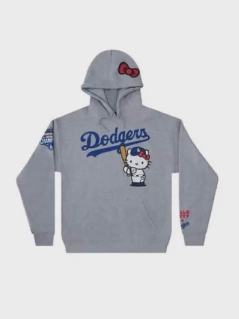 Op de afbeelding: Grijze hoodie met het woord "Dodgers" in blauw script en een Hello Kitty-personage met een honkbalknuppel. De capuchon heeft een rode strik. Op de mouwen zitten patches met tekst. Een kangoeroezak is zichtbaar.