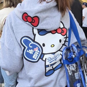 Op de afbeelding: Grijze hoodie met een grote Hello Kitty-afbeelding in wit, rood en blauw, met een blauw-witte foam finger met de tekst "LA" en "Hello Kitty". De hoodie heeft ook het Dodgers-logo en een rode strik op de capuchon.