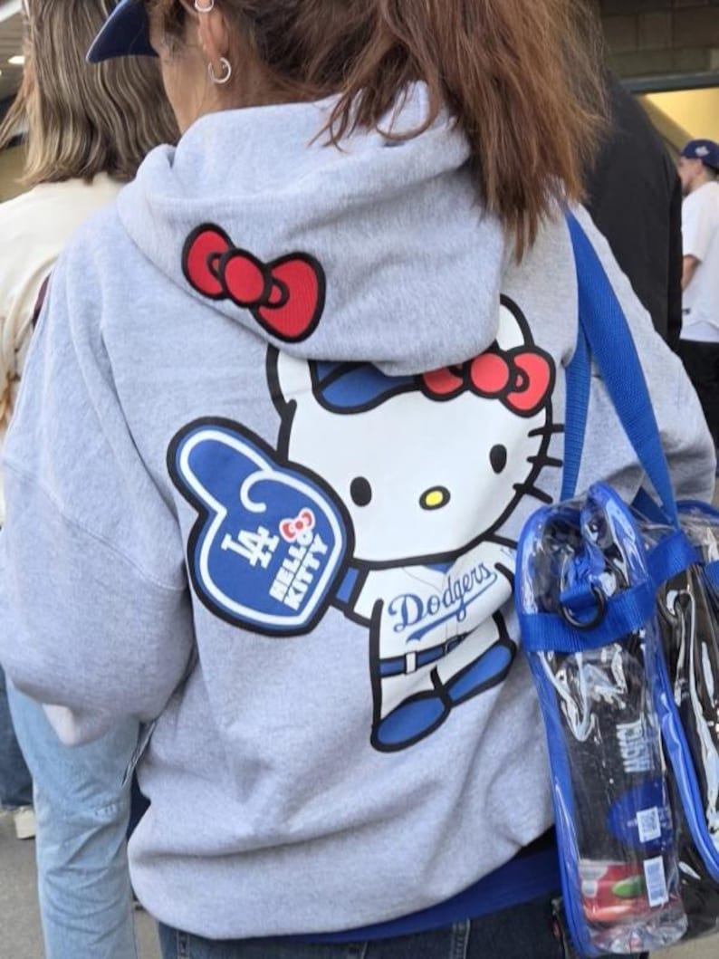 Op de afbeelding: Grijze hoodie met een grote Hello Kitty-afbeelding in wit, rood en blauw, met een blauw-witte foam finger met de tekst "LA" en "Hello Kitty". De hoodie heeft ook het Dodgers-logo en een rode strik op de capuchon.