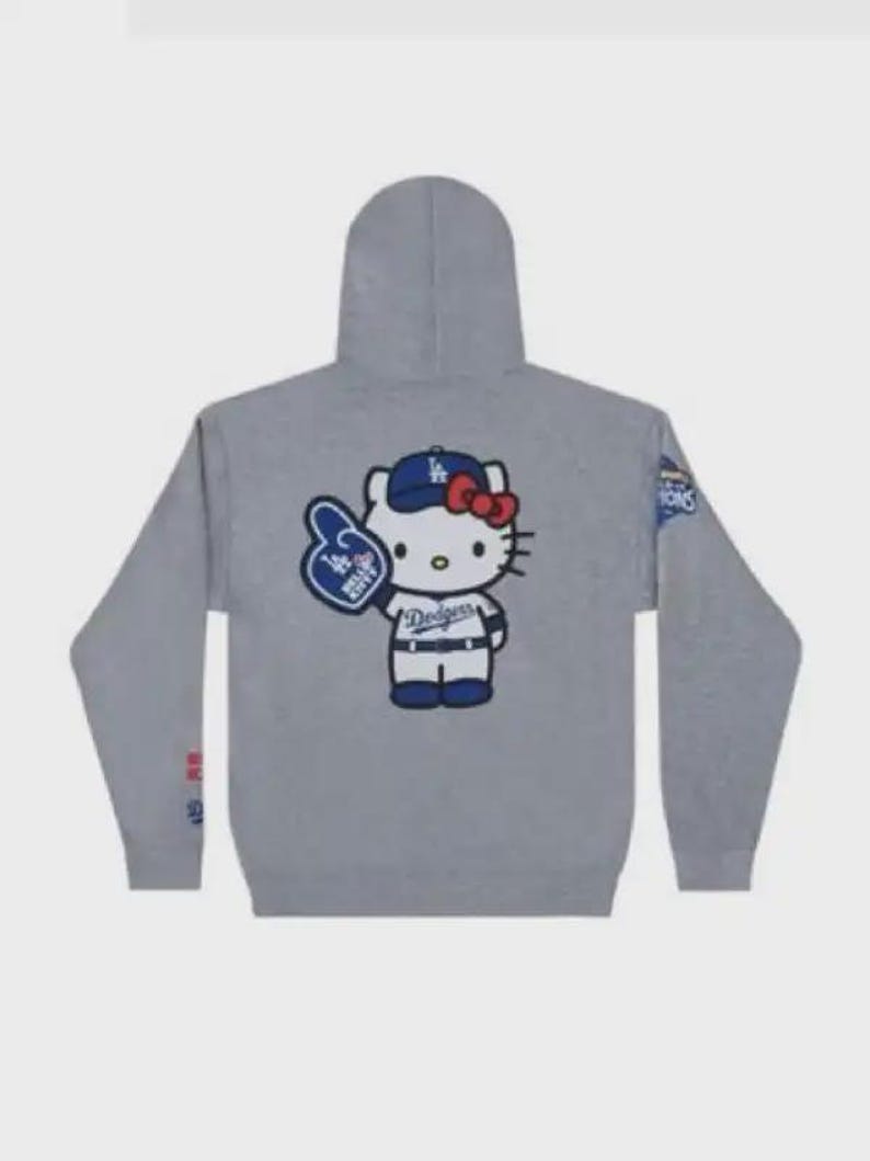 Op de afbeelding: Grijze hoodie met een Hello Kitty-afbeelding met een Dodgers-baseballpet en een foam finger. De hoodie heeft een capuchon en lange mouwen. Het Dodgers-logo is zichtbaar.