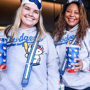 Op de afbeelding: Twee vrouwen dragen grijze hoodies met het Los Angeles Dodgers-logo en een Hello Kitty-afbeelding. Ze dragen ook Hello Kitty-hoofdbanden en dragen bijpassende bekers en tassen. De tassen hebben blauw-witte gestreepte bandjes.