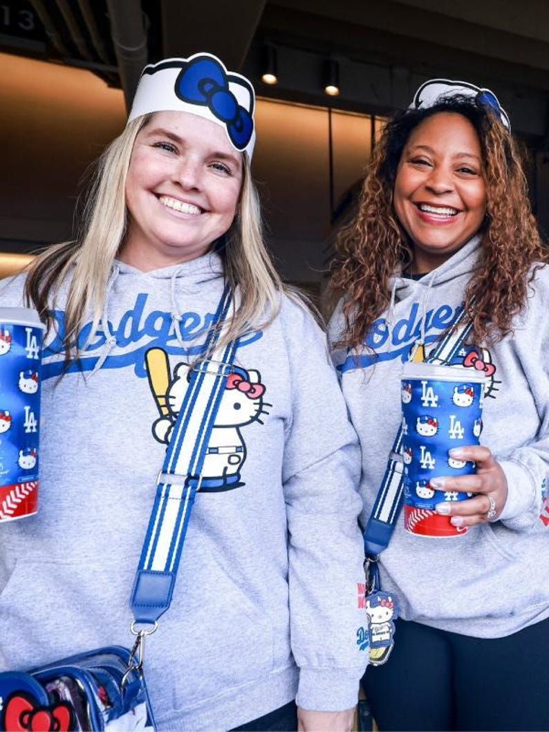 Op de afbeelding: Twee vrouwen dragen grijze hoodies met het Los Angeles Dodgers-logo en een Hello Kitty-afbeelding. Ze dragen ook Hello Kitty-hoofdbanden en dragen bijpassende bekers en tassen. De tassen hebben blauw-witte gestreepte bandjes.