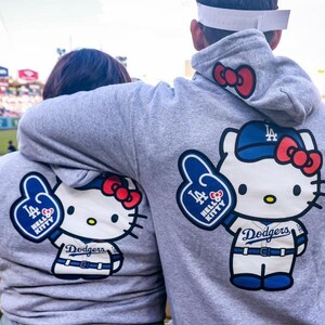 Op de afbeelding: Twee grijze hoodies met een Hello Kitty-ontwerp in een Dodgers honkbaluniform. Het ontwerp omvat een blauwe honkbalpet, een rode strik en een foam finger. De hoodies hebben een rode strik op de capuchon.