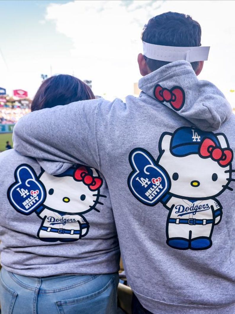 Op de afbeelding: Twee grijze hoodies met een Hello Kitty-ontwerp in een Dodgers honkbaluniform. Het ontwerp omvat een blauwe honkbalpet, een rode strik en een foam finger. De hoodies hebben een rode strik op de capuchon.
