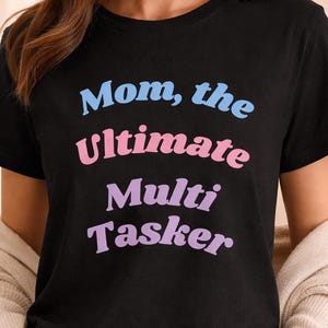 Puede incluir: Camiseta negra con el texto "Mom, the Ultimate Multi Tasker" en azul, rosa y morado. La camiseta se combina con un pantalón de chándal gris y una rebeca beige. Un atuendo casual y cómodo.