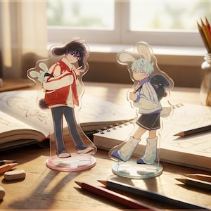 May include: Two acrylic anime character figures with bunny ears stand on a desk with open sketchbooks, pencils, and erasers. One figure wears a red jacket, the other a white jacket. The scene is lit by natural light, suggesting a creative workspace.