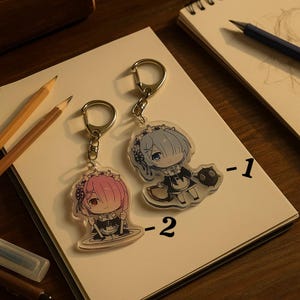 May include: Two anime-style acrylic keychains with silver-colored key rings. One keychain features a character with blue hair, and the other has pink hair. Both characters are wearing black and white outfits.